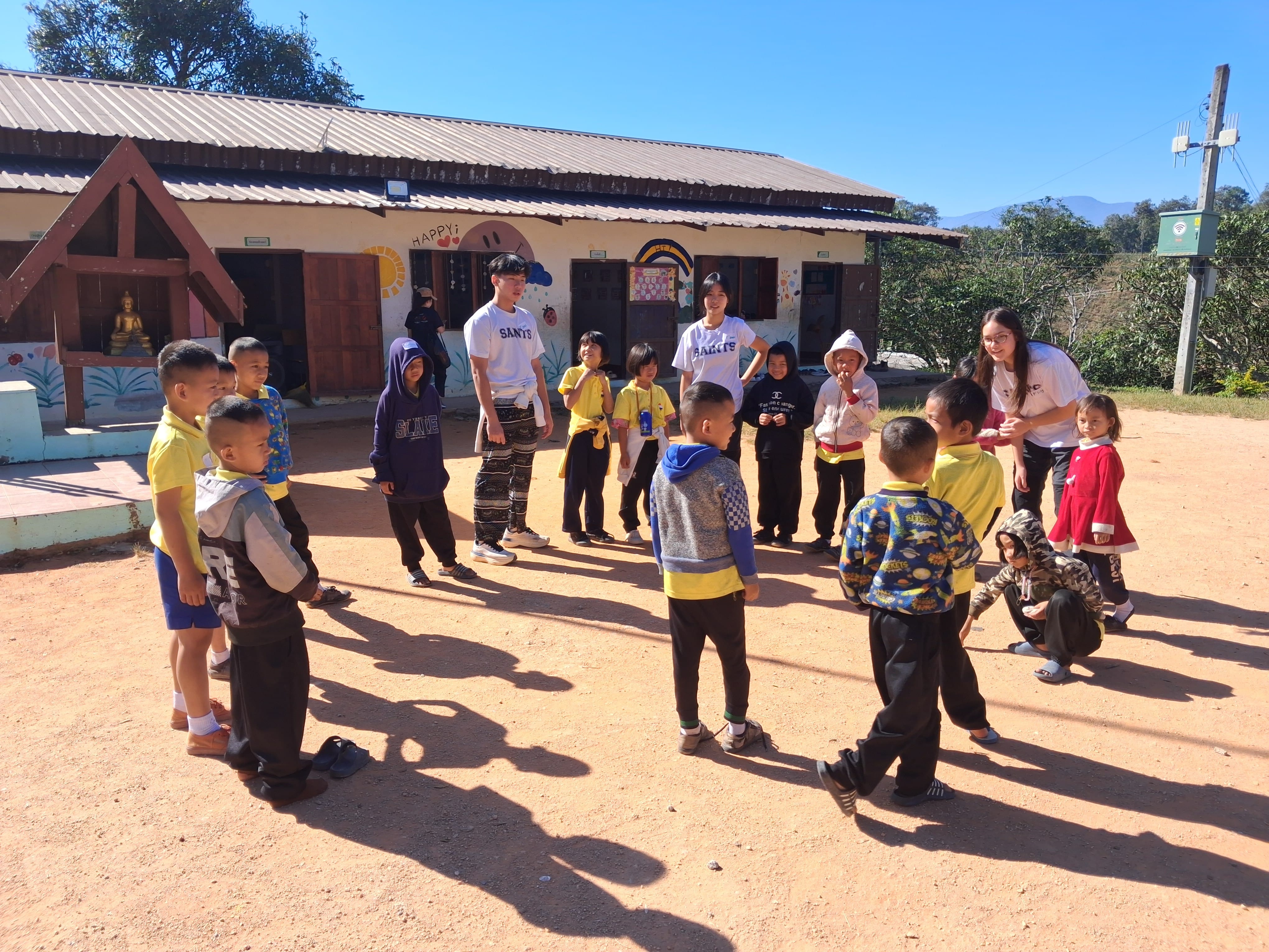 Student volunteers from wearing SAINTS t-shirts playing an outdoor circle game with young school children on a dirt playground in front of a rural school building under a clear blue sky.
